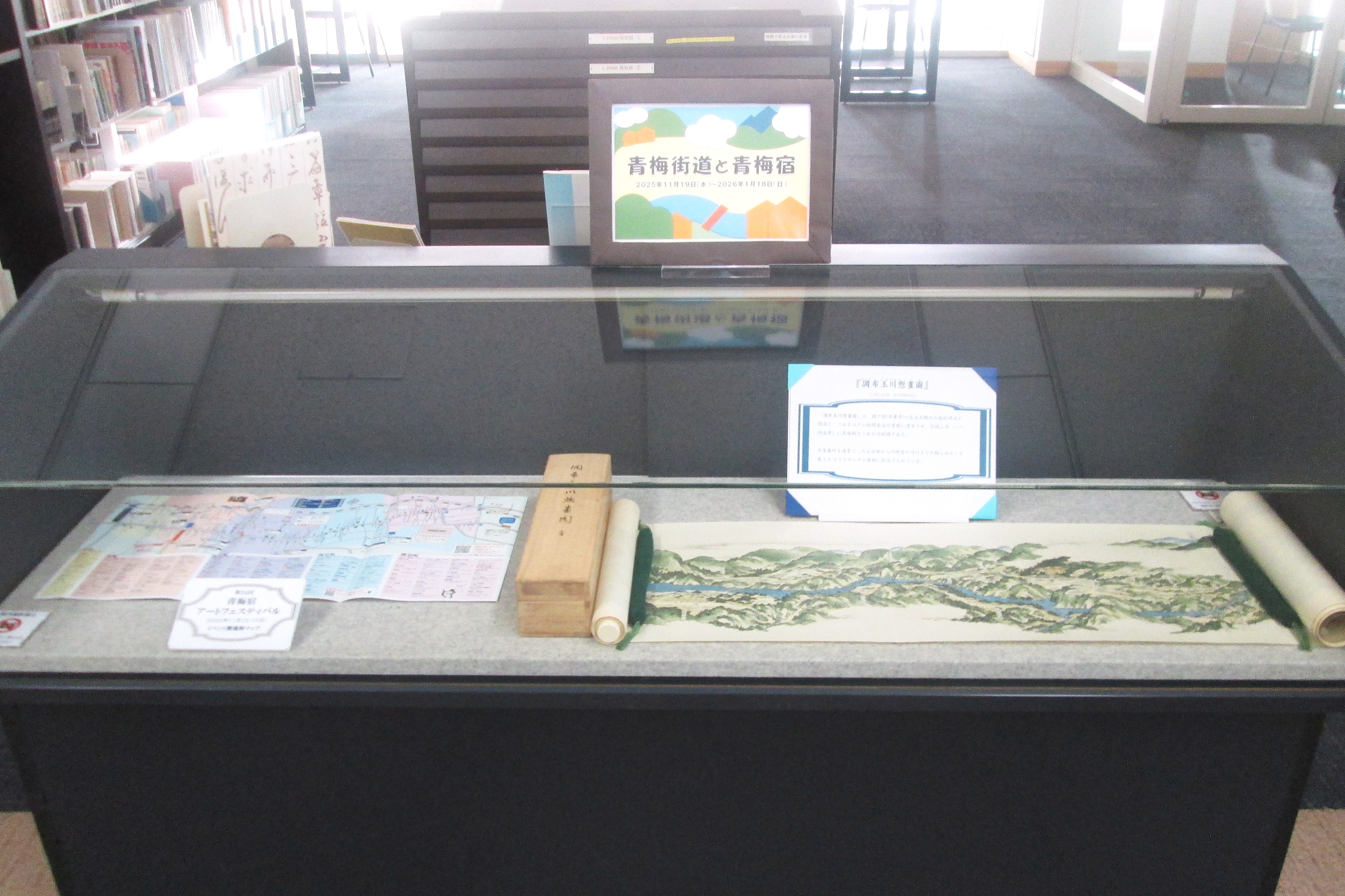 横長のガラスケースが写っている写真。
ガラスケース内左側に青梅宿アートフェスティバルのリーフレット、右側に調布多摩川惣画図がおさめられている。